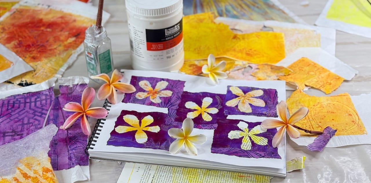 Flower artwork made from torn paper collage. A paper painting. . Top down view of artwork surrounded by collage papers, gel prints, frangipani / plumeria flowers, gloss medium collaging glue and a brush in a pot.