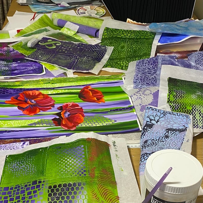 a painting of 3 poppies in the process of being collaged. the painting is surrounded by gel prints. The prominent colours are green and purple. The poppies are red.