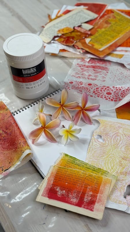 Top down view of art book surrounded by collage papers, gel prints, frangipani / plumeria flowers, and gloss medium collaging glue