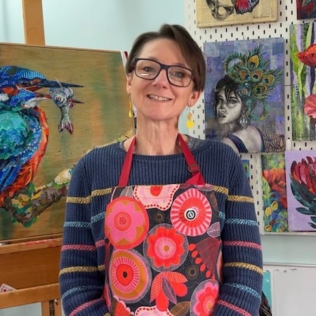 A lady, Amber Bennison, standing in front of an easel surrounded by her collage art. The lady is wearing a bright apron. The pominant artworks are of a kingfisher, a portrait and some flowers.