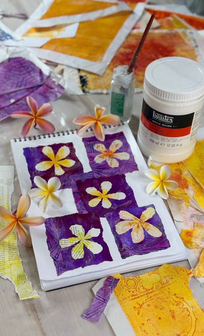 Flower artwork made from torn paper collage. A paper painting. . Top down view of artwork surrounded by collage papers, gel prints, frangipani / plumeria flowers, gloss medium collaging glue and a brush in a pot.