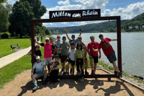 People standing under "Mitten am Rhein" sign next to the Rhine River. CRAP H3 Hash House Harriers - Cologne, Bonn, Düsseldorf