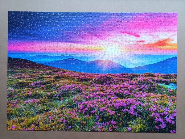 Bild von einem Puzzle mit buntem Himmel und einer Blumenwiese vor einer Bergkulisse.