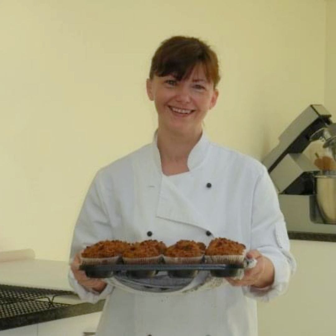 Sharon Burchall, founder of Sharon Burchall Cake Studio — smiling and holding a tray of freshly baked muffins, known in New Zealand as “The Muffin Lady” before becoming a cake business owner and cake decorating teacher.