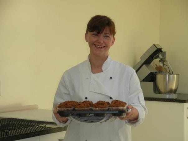 Sharon Burchall, founder of Sharon Burchall Cake Studio — smiling and holding a tray of freshly baked muffins, known in New Zealand as “The Muffin Lady” before becoming a cake business owner and cake decorating teacher.