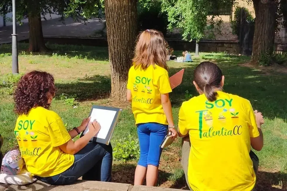 Personas con camisetas amarillas de TalentiaCC realizando actividades educativas al aire libre, dibujando y observando la naturaleza en un parque