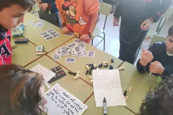 Niños y niñas participando en una actividad educativa de lógica y resolución de acertijos en el aula en un taller de TalentiaEx, con piezas de ajedrez, cartas y materiales didácticos sobre mesas escolares