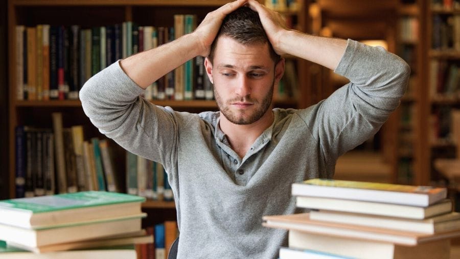 Man met handen op hoofd die vastloopt tussen stapels boeken, symbool voor uitstelgedrag bij ADHD