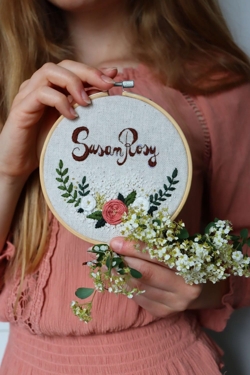 Žena v ružovej blúze držiaca v rukách okrúhlu výšivku plnú kvetov, ruží, lístkov a nápisom SusanRosy, v svetlom bambusovom ráme, vedľa výšivky sú biele drobné čerstvé kvety.