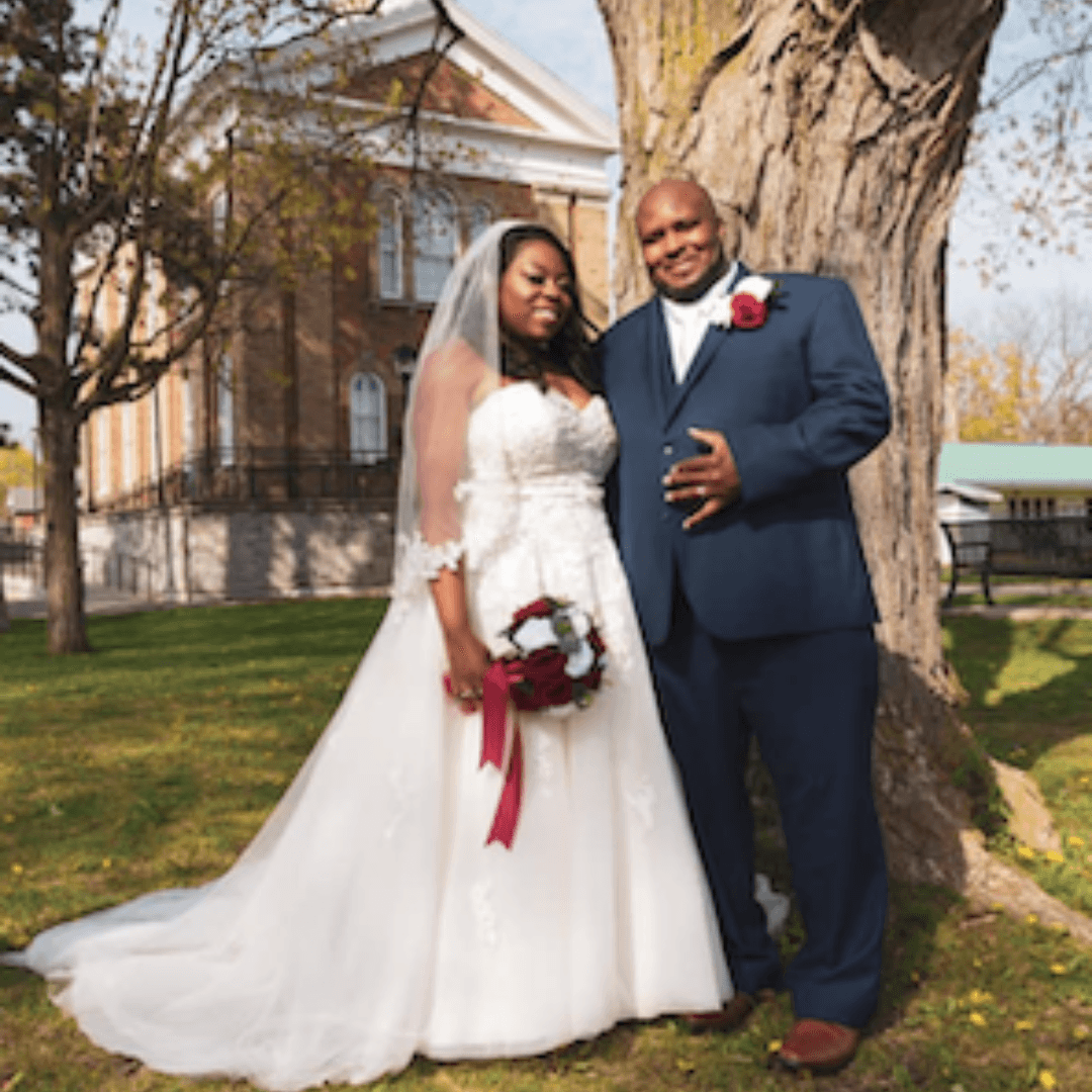 Bride in a white lace gown holding a bouquet and standing with her groom beside a large tree on their wedding day.