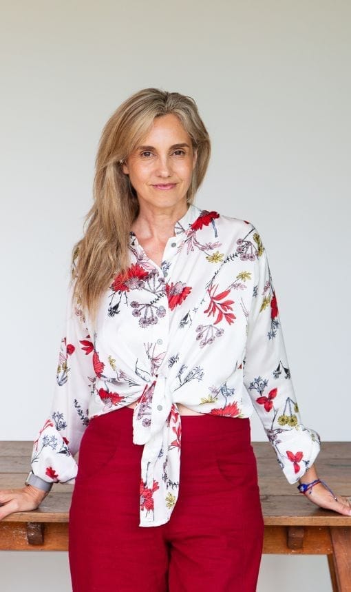 Karina Neira de pie con blusa de flores y pantalón rojo, sonriendo en un entorno luminoso.