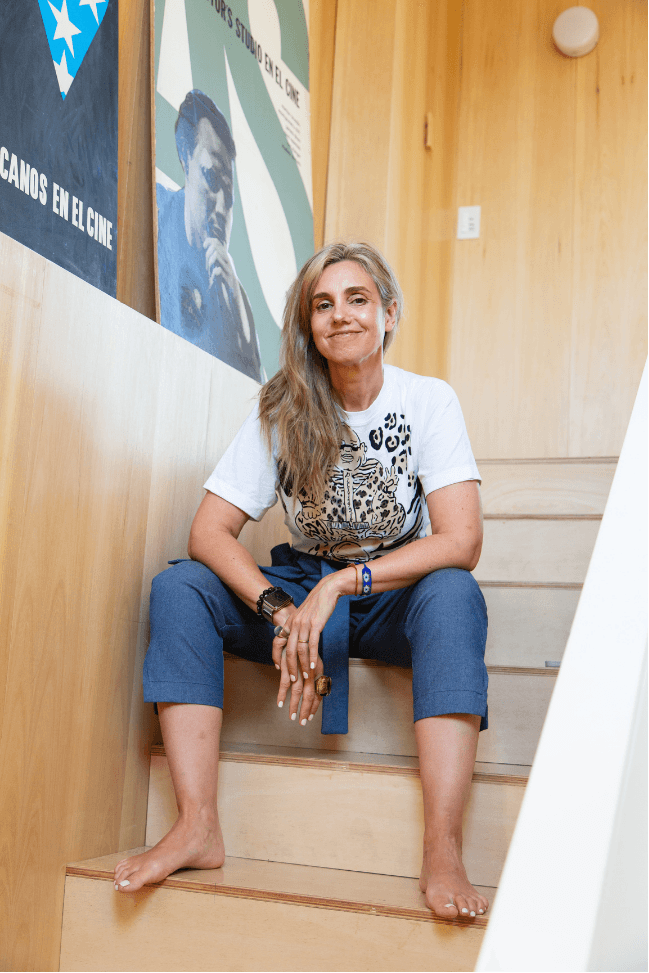 Kari Neira descalza sentada en una escalera de madera, luciendo camiseta con estampado de leopardo y pantalón azul, sonriendo.