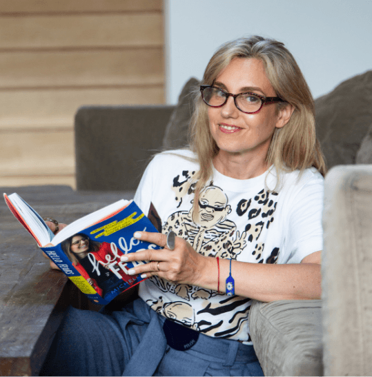 Kari Neira con gafas leyendo un libro de tapa roja en un sofá; viste camiseta con ilustración
