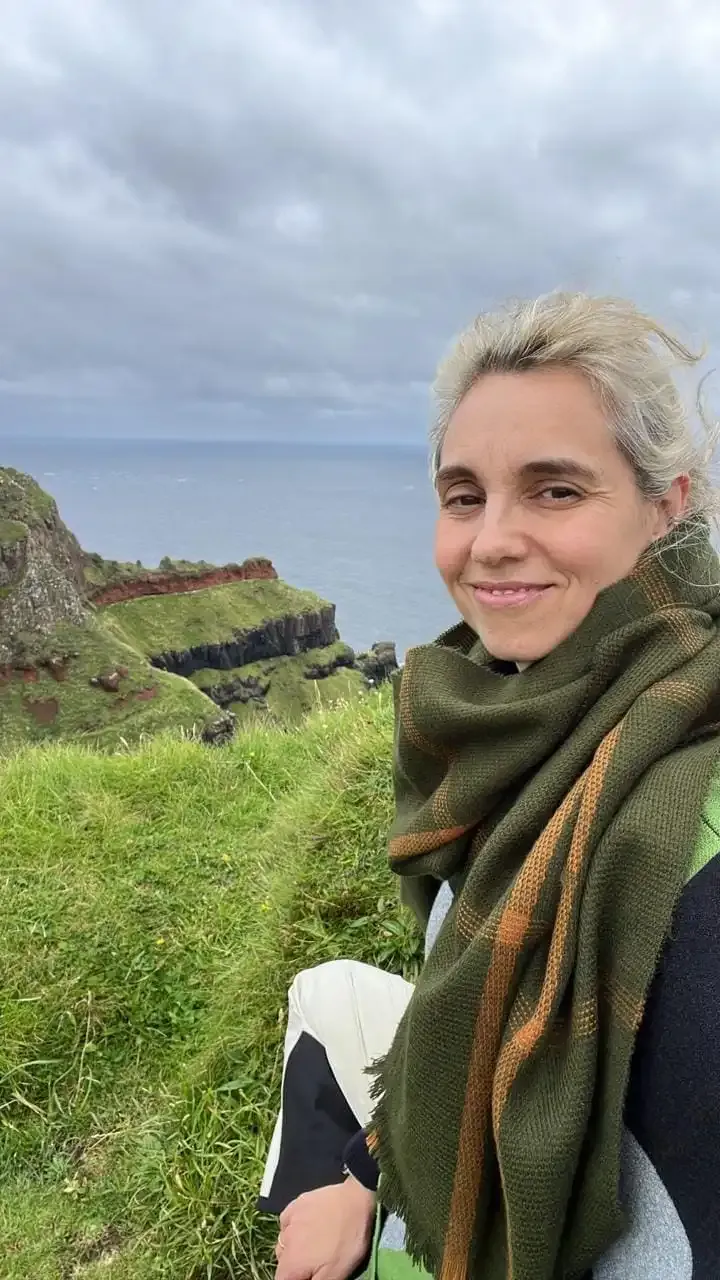 Kari Neira con bufanda verde y chaqueta negra sonriendo, sentada frente a un acantilado verde con mar al fondo”