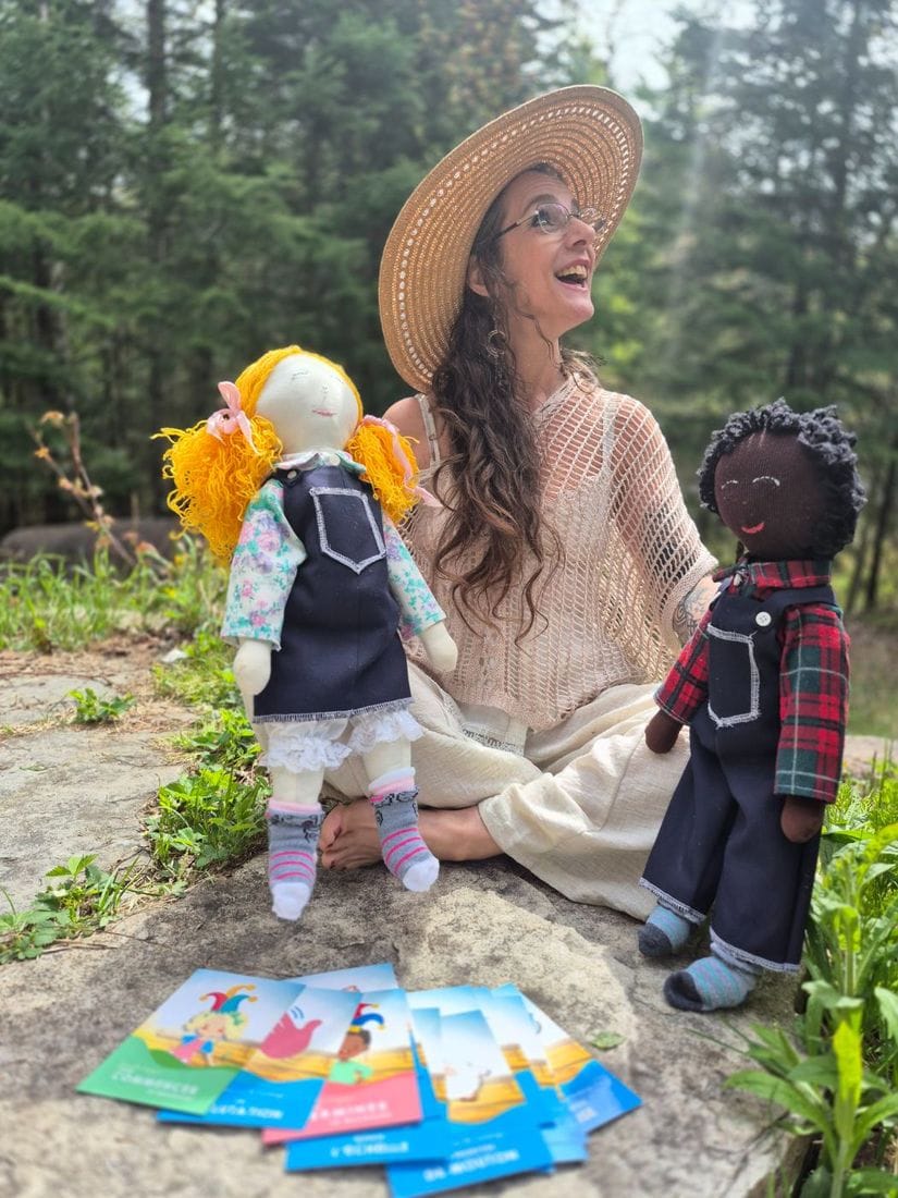 Annie Laberge assise sur une roche en forêt, les poupées Guillaume et Charlotte à ses côtés et les cartes de massage en main.