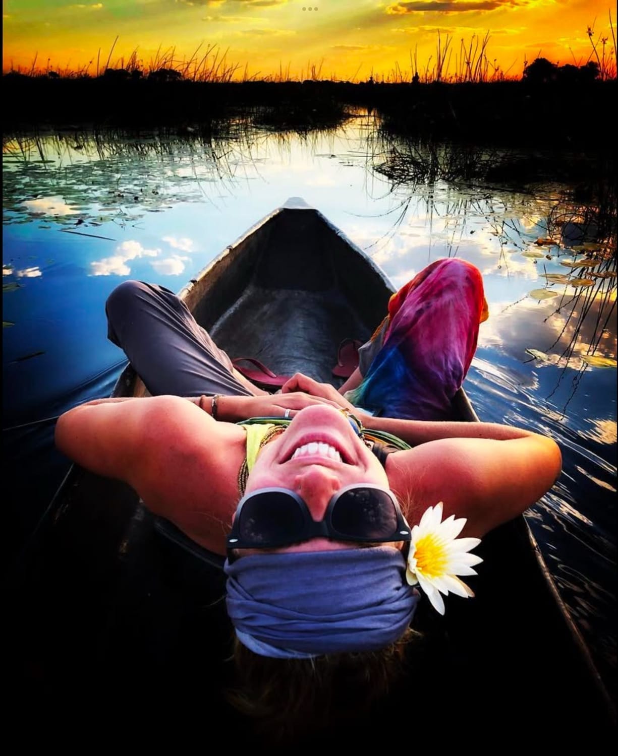 Danielle Blum smiles brightly while reclining in a carved-out wooden canoe in Botswana, wearing sunglasses, a headscarf, and a flower tucked behind her ear. The sunset sky glows in shades of gold and orange, reflecting across the still water and surrounding reeds. A vibrant, soulful travel moment capturing the beauty of Africa and the joy of adventure.