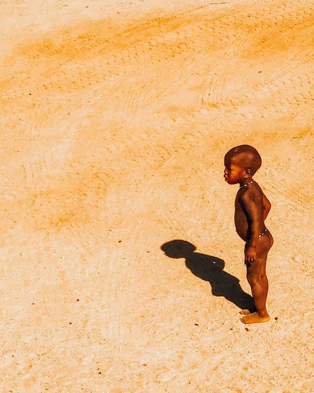 Young child standing barefoot on sunlit sand in rural Africa, casting a long shadow—an evocative moment reflecting soulful travel, cultural immersion, and bespoke journeys curated by a boutique travel planner.