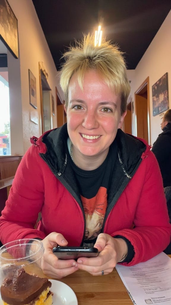 Sassi sitzt lächelnd in einem Café, trägt eine rote Jacke und hält ihr Handy in der Hand. Vor ihr steht ein Stück Kuchen und ein Getränk auf dem Holztisch – eine entspannte und sympathische Szene.