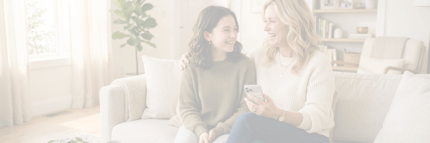 Blonde Frau Mitte 50 sitzt mit ihrer Teenagertochter auf dem Sofa, beide lächeln sich warm an, Mutter hält ein Smartphone, helles freundliches Wohnzimmer, geborgene und liebevolle Atmosphäre