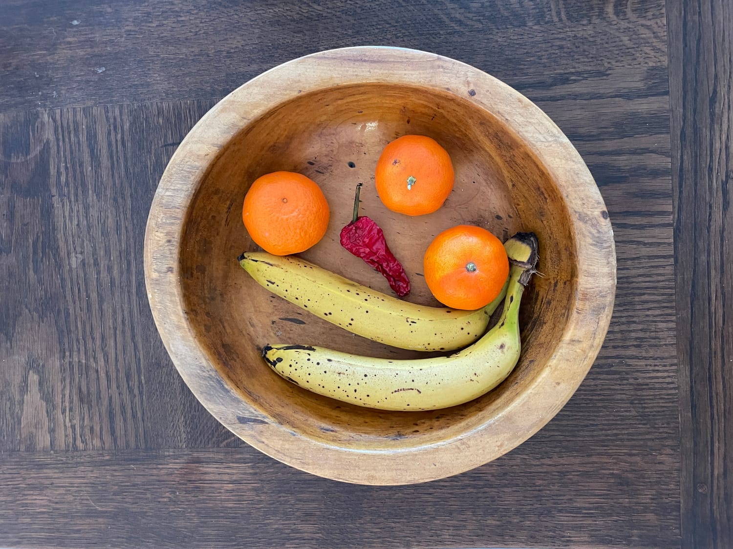 Hölzerne Obstschale auf einem dunklen Holztisch. Darin zwei gelbe Bananen mit braunen Punkten, drei kleine Mandarinen und eine getrocknete rote Chili. Zwischen den runden Früchten wirkt die Chili wie ein kleiner Aufstand im Obstkorb. Alttextpoesie