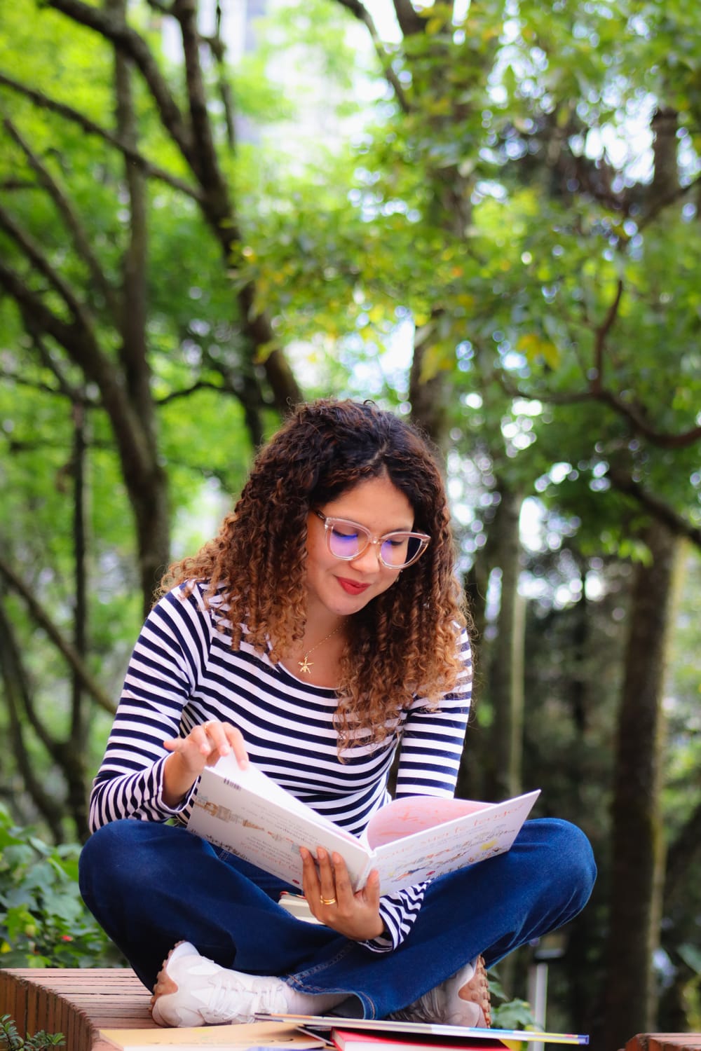 Maria Camila Tafur sentada en un parque leyendo un libro infantil, usa camiseta de rayas, el cabello suelto y crespo y gafas. Sonríe levemente. 