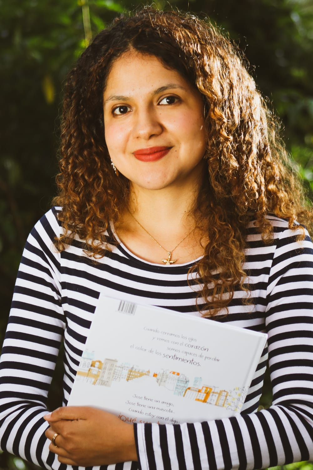 Maria Camila Tafur mira hacia la cámara, con una leve sonrisa, su cabello crespo es ilumindao por el sol. Tiene un libro en la mano y usa una camiseta de rayas.