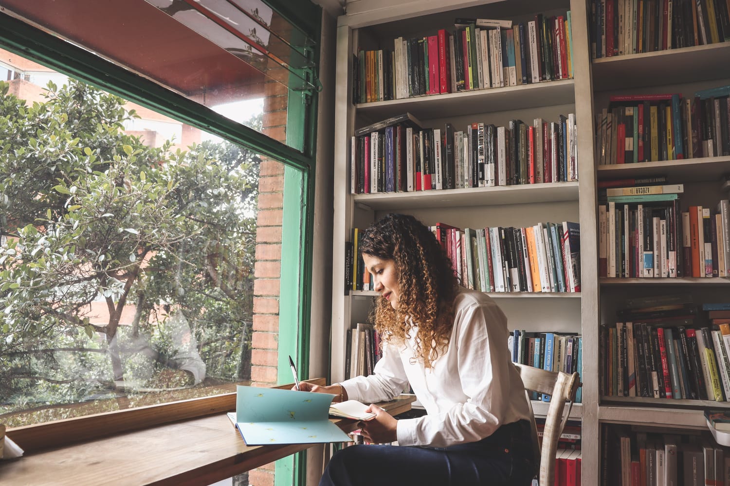 Maria Camila Tafur en una librería, está sentada revisando un libro infantil y tomando notas en su libreta. Usa camisa blanca y jean. Si cabello es dorado y crespo. 