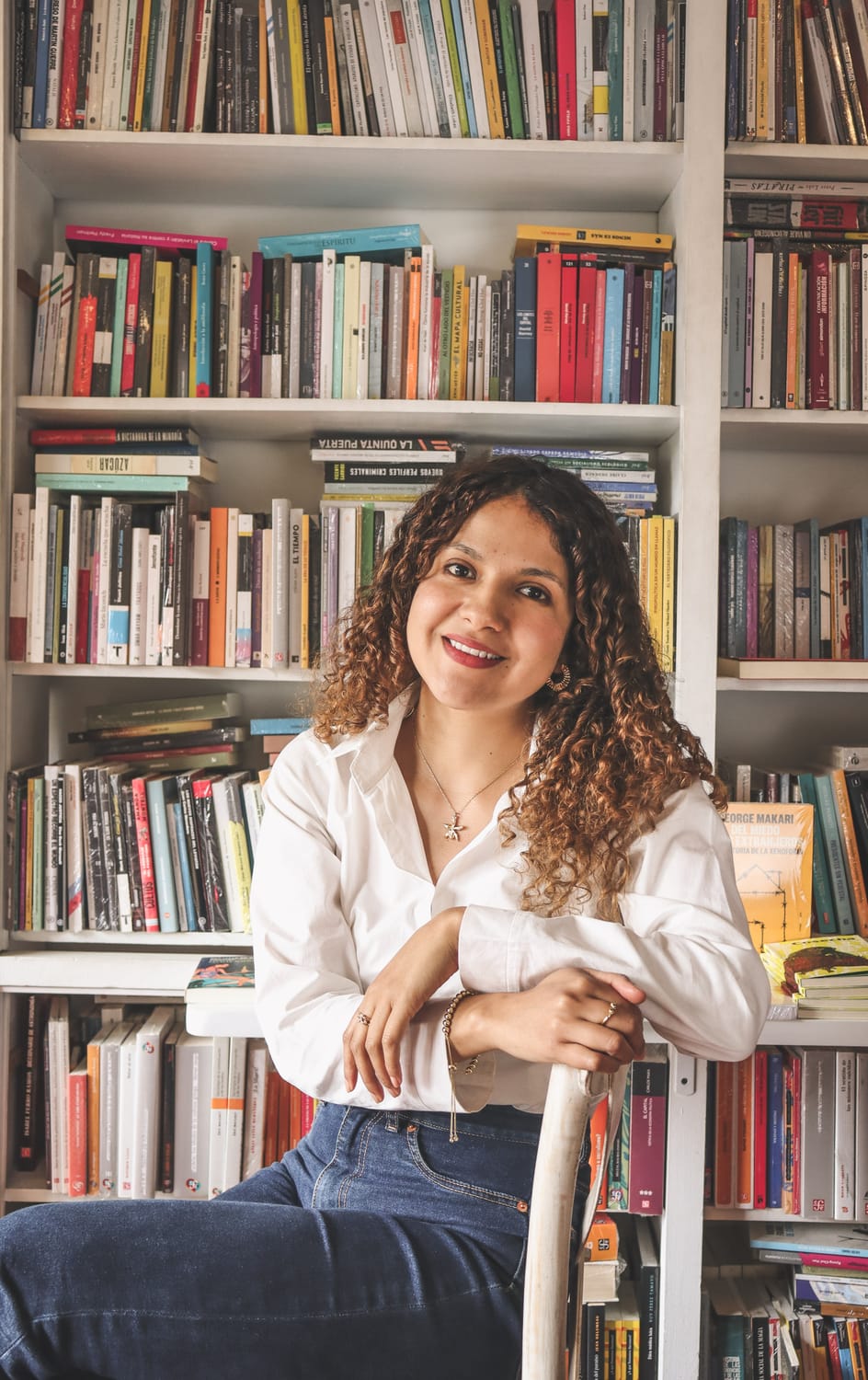 Maria Camila Tafur sentada en una silla blanca usa camisa blanca y jean oscuro. Su cabello es crespo y dorado, sonríe mientras mira a la cámara, atrás de ella hay una biblioteca con libros de diversos tamaños y colores.