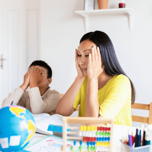 Frustrated mother and child sitting at a table with school materials, both covering their faces with their hands, overwhelmed during homeschooling.
