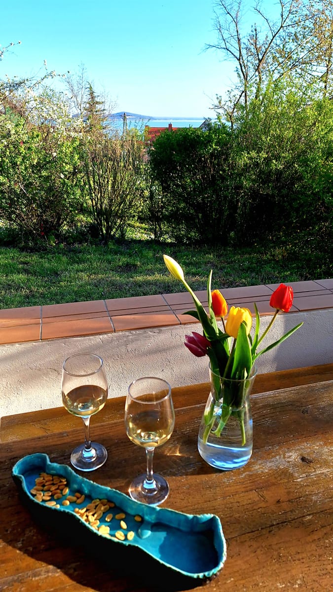 Asztal a verandán, két pohár borral, tulipánokkal és Balaton alakú kerámiatállal, háttérben balatoni kilátással Badacsonytomajon