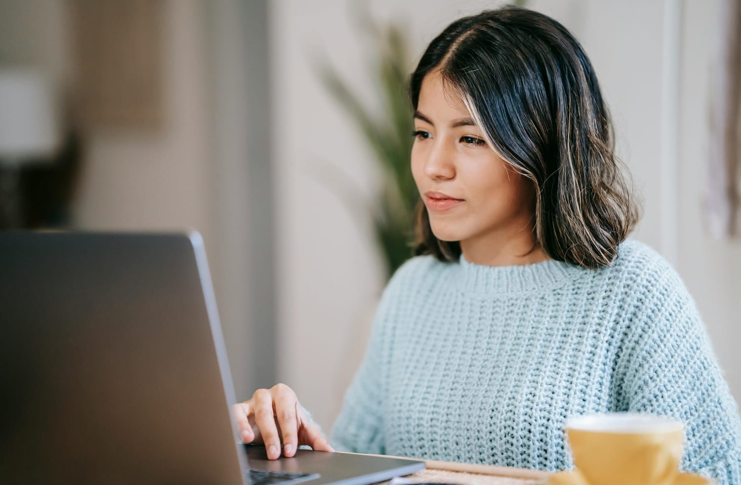 woman in blue sweater with dark hair and highlights working on a laptop with a coffee cup in front of her