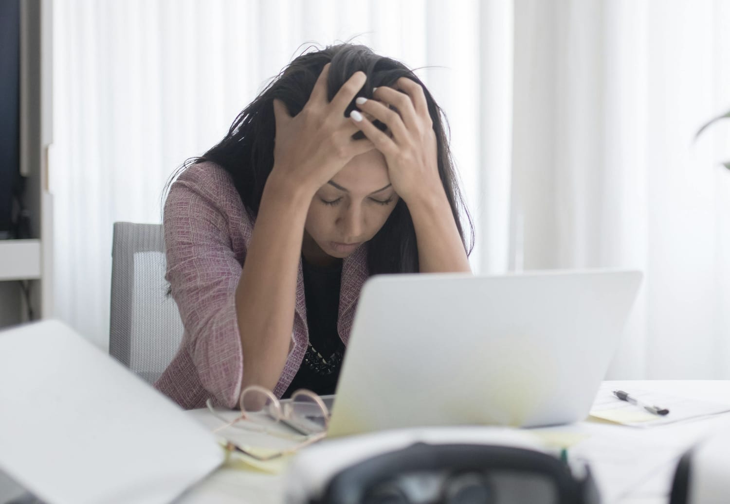 A woman sitting at a cluttered desk with her head in her hands, looking overwhelmed while staring at her laptop.