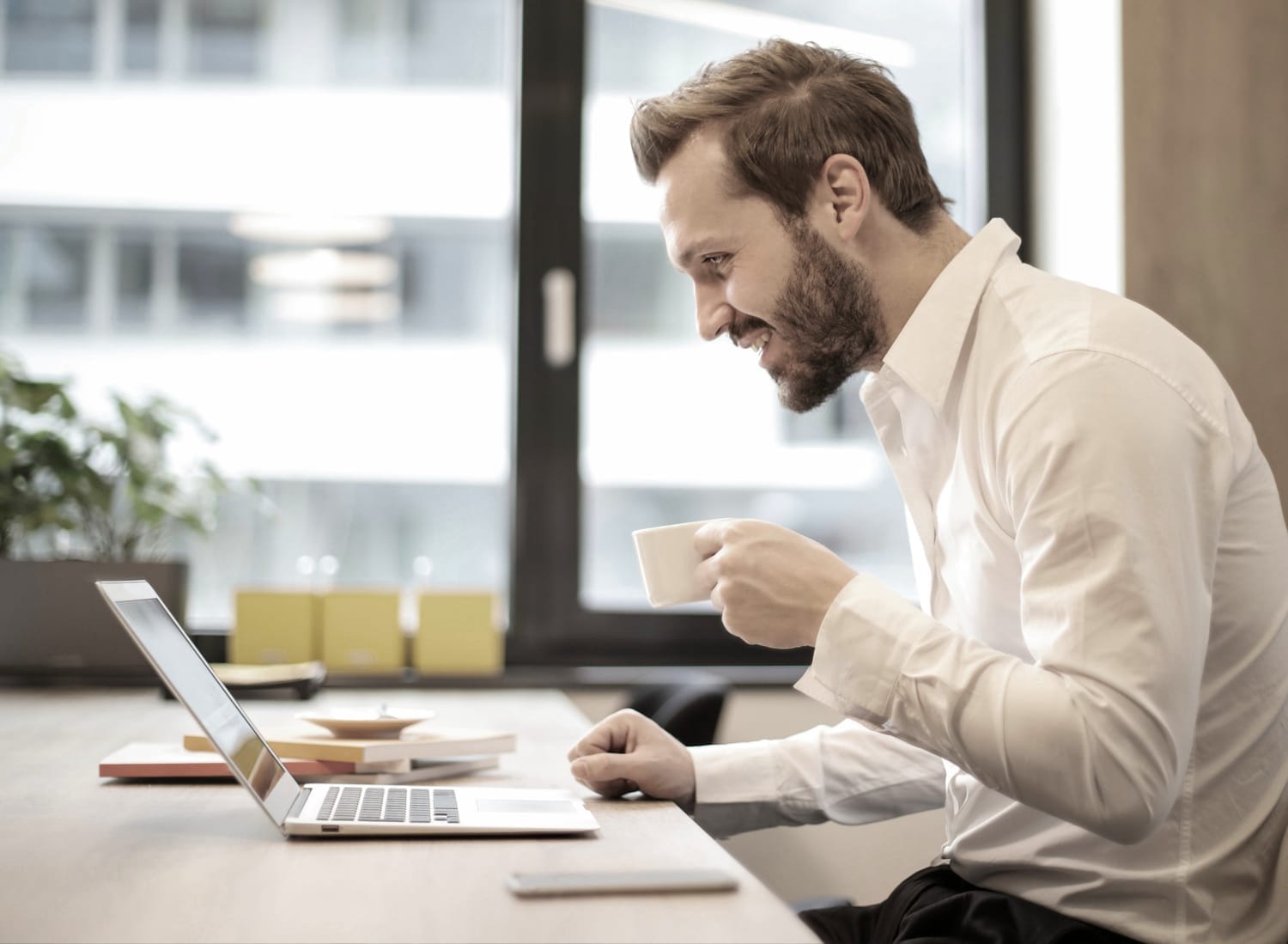 A man smiling while working on a laptop at a clean desk, holding a coffee cup and appearing focused and confident.