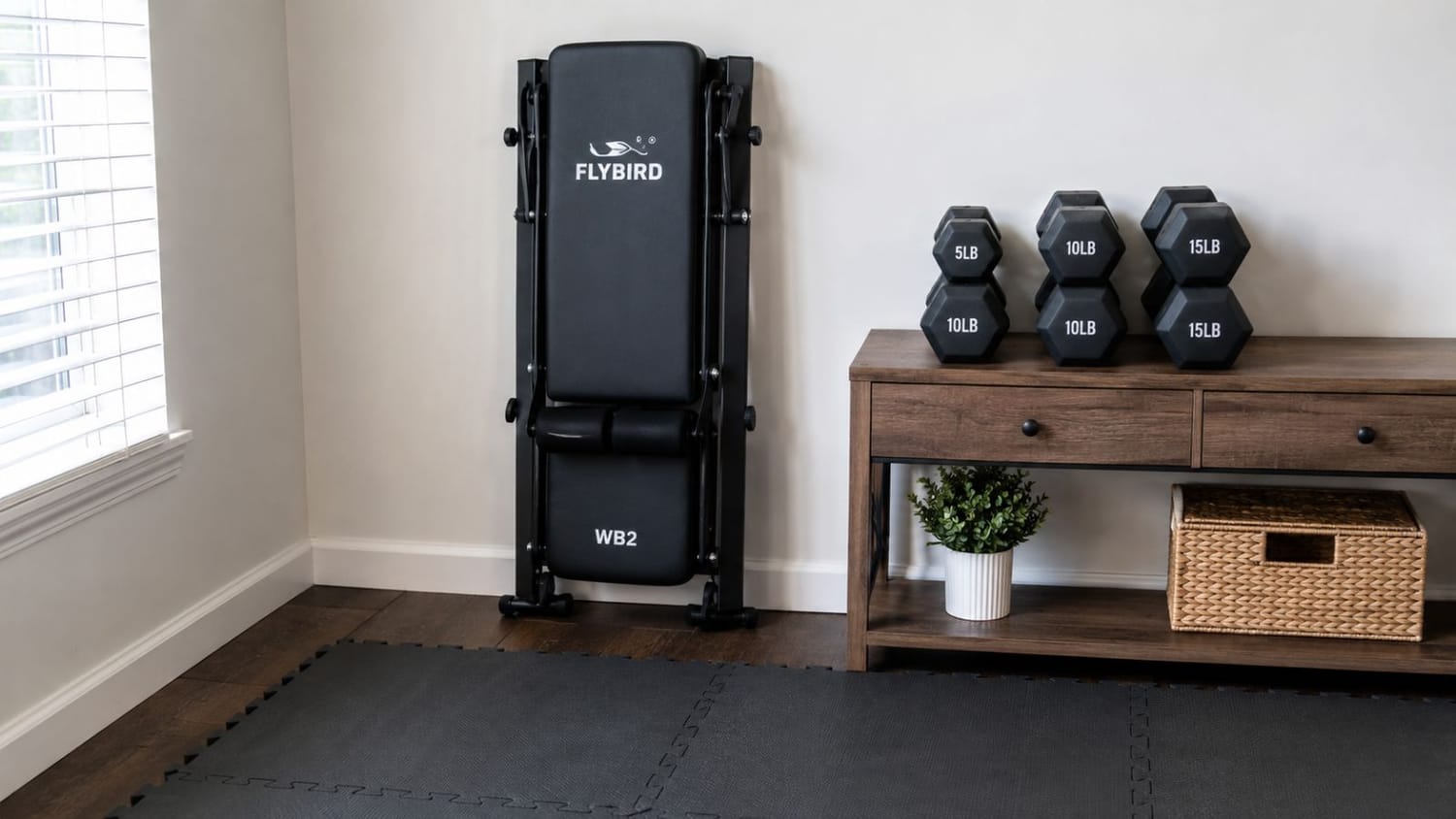 Folded adjustable weight bench leaning against a neutral apartment wall next to dumbbells