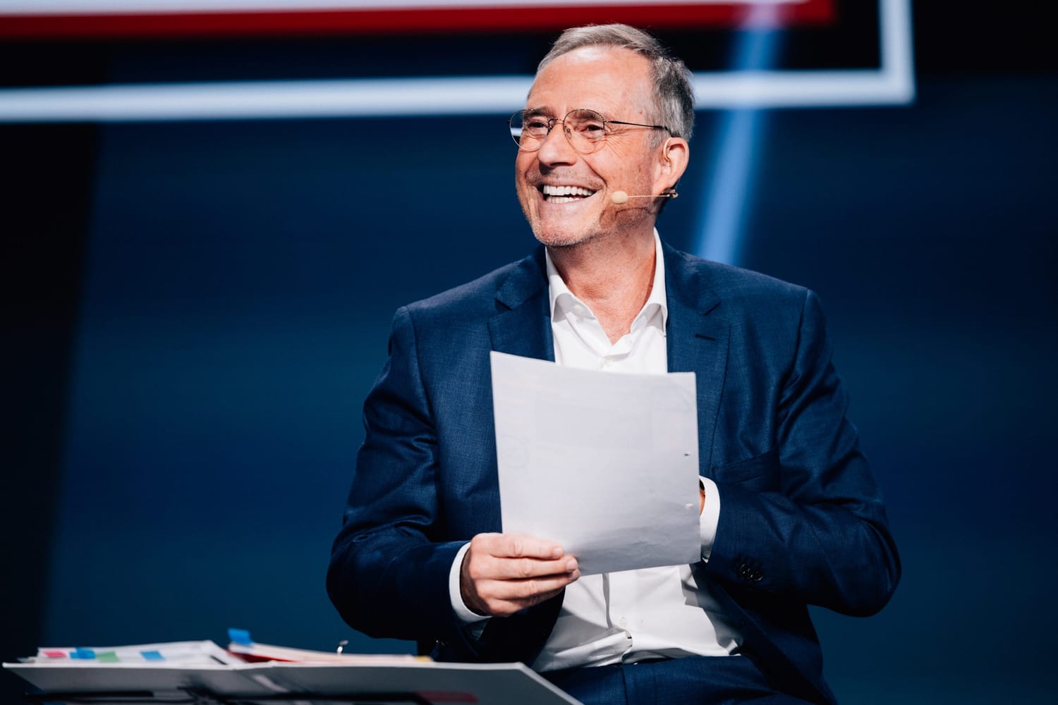 Bodo Schäfer sitzt auf einer Bühne, trägt einen dunklen Anzug und hält ein Blatt Papier in der Hand. Er lächelt und wirkt entspannt, während er mit einem Headset-Mikrofon spricht. Im Hintergrund ist eine moderne Bühnenkulisse mit dunklen Farben und Lichtakzenten zu sehen.