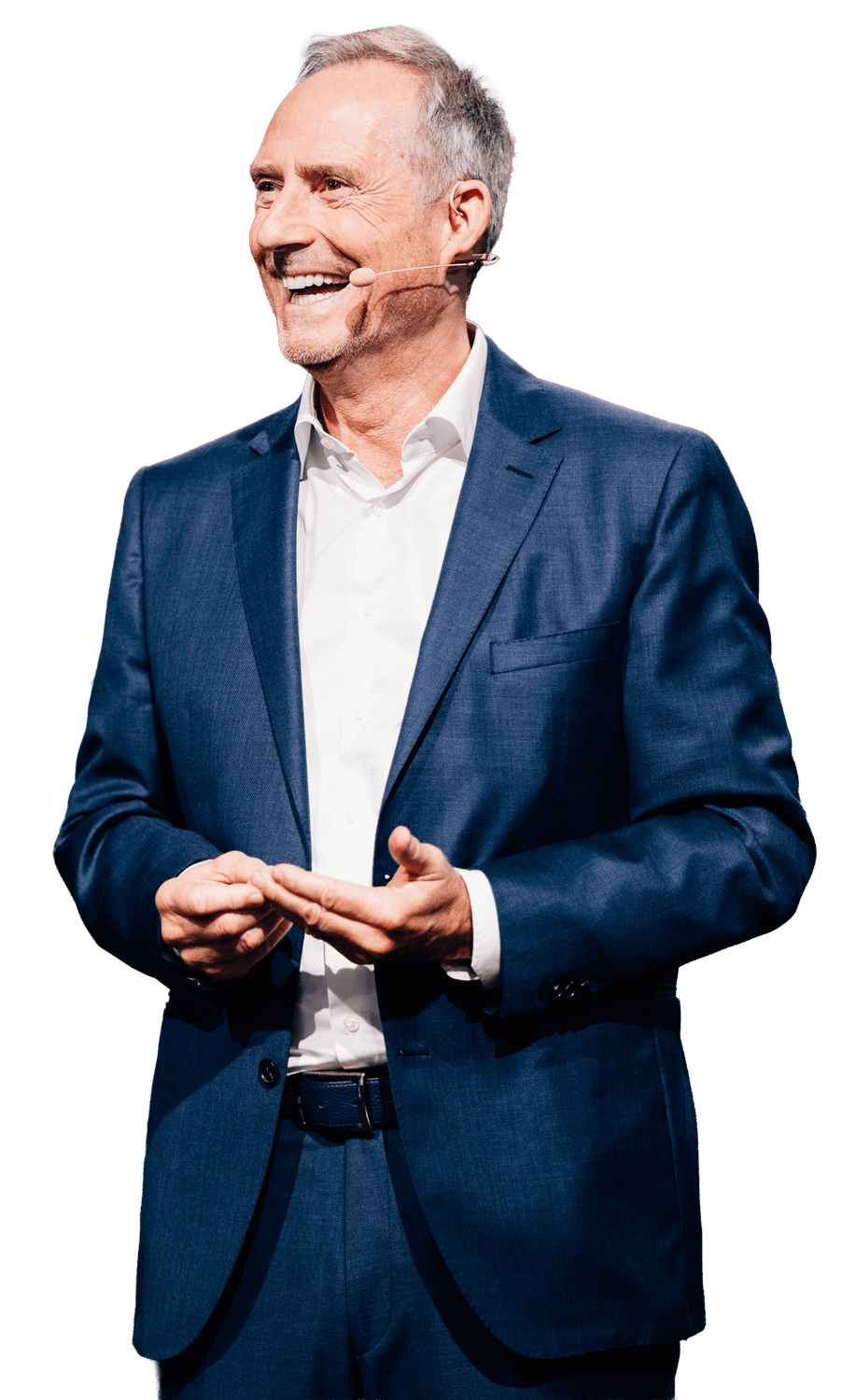 Bodo Schäfer steht vor dunklem Hintergrund und trägt einen eleganten Anzug mit weißem Hemd. Er lächelt und wirkt locker, während er mit einem Headset-Mikrofon spricht. Seine Hände sind leicht nach vorne gerichtet, als würde er gerade etwas erklären oder einen wichtigen Punkt unterstreichen.