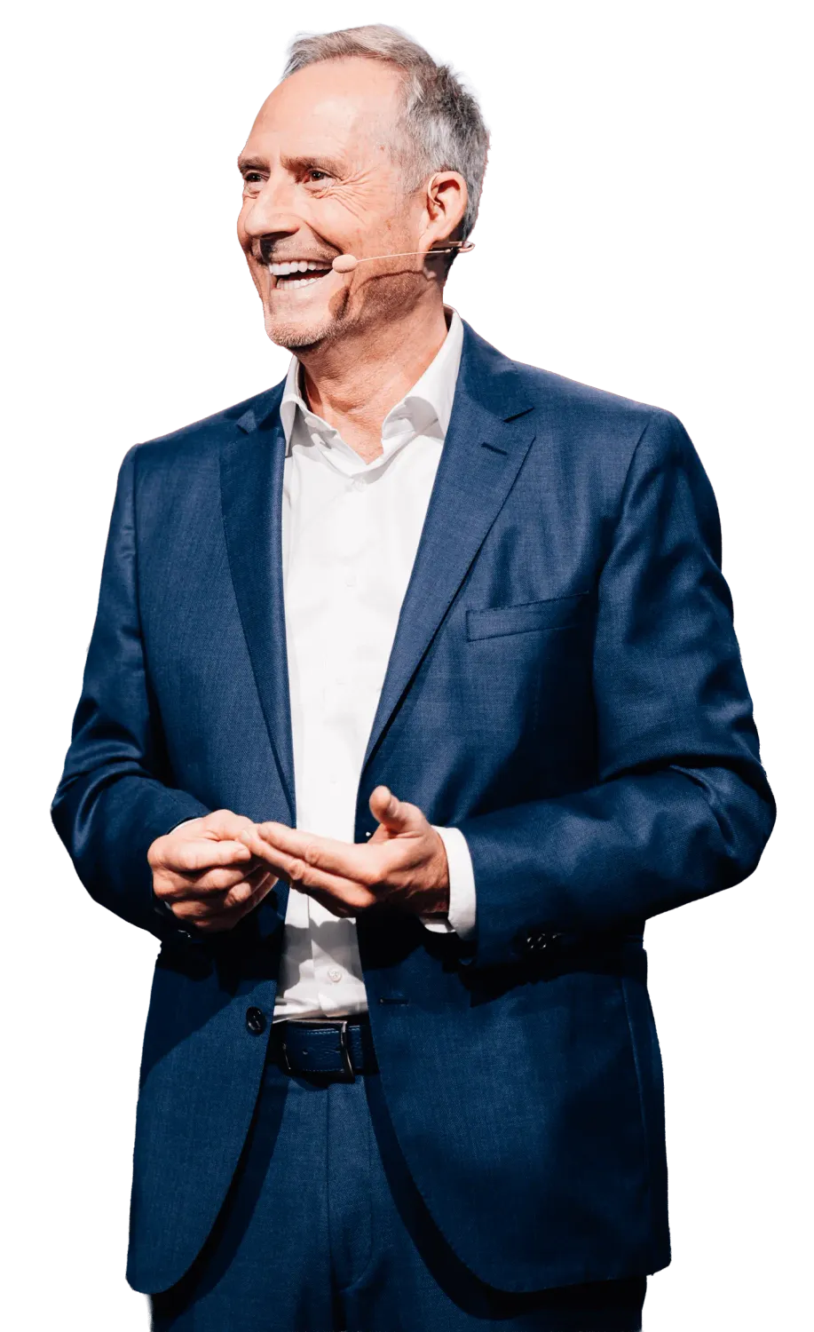 Bodo Schäfer steht vor dunklem Hintergrund und trägt einen eleganten Anzug mit weißem Hemd. Er lächelt und wirkt locker, während er mit einem Headset-Mikrofon spricht. Seine Hände sind leicht nach vorne gerichtet, als würde er gerade etwas erklären oder einen wichtigen Punkt unterstreichen.