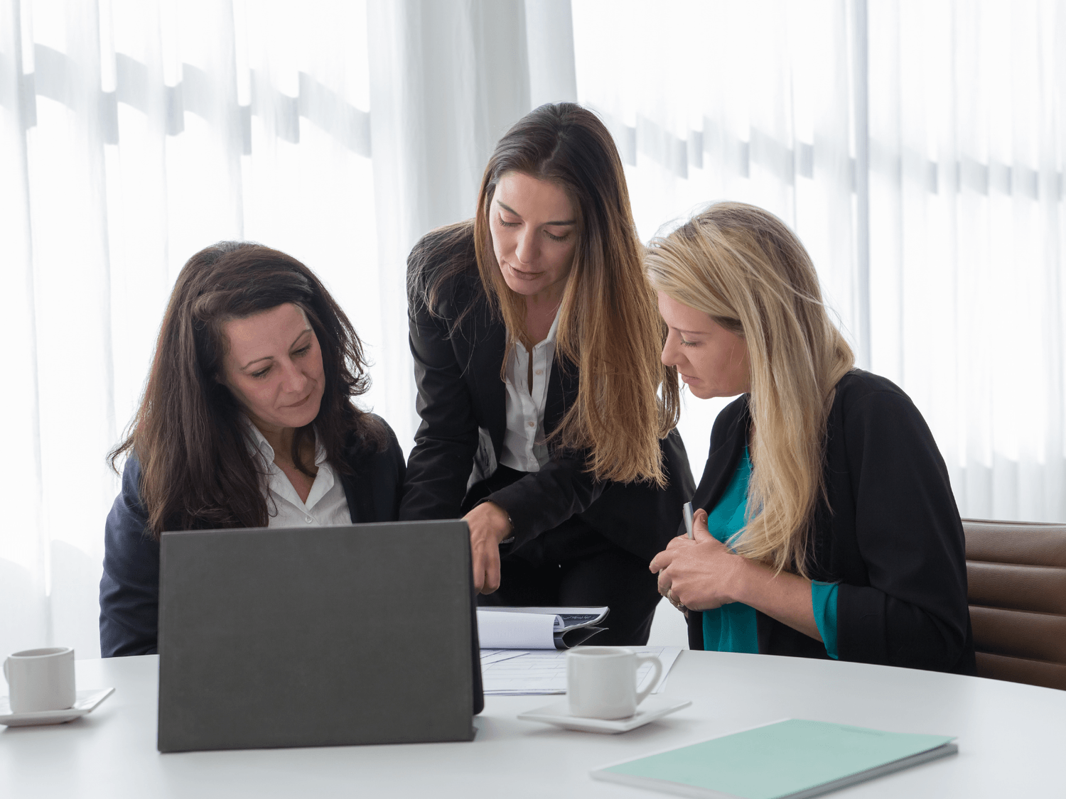 „Trzy kobiety w biznesowych stylizacjach dyskutują przy stoliku nad notatkami i laptopem podczas spotkania firmowego.