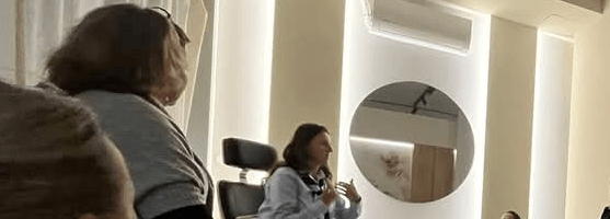 Uczestniczka warsztatu Kobiety 100/100 słucha prelekcji, a prowadząca Jolanta Valentin widoczna jest w tle.”