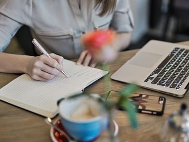 Kobieta pisząca w notesie przy stoliku, z otwartym laptopem i odłożonym telefonem, sfotografowana przez rozmyty kwiat.