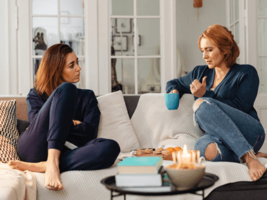 Dwie kobiety siedzą na kanapie i rozmawiają; jedna trzyma filiżankę, przed nimi stoi stolik z książkami i przekąskami.