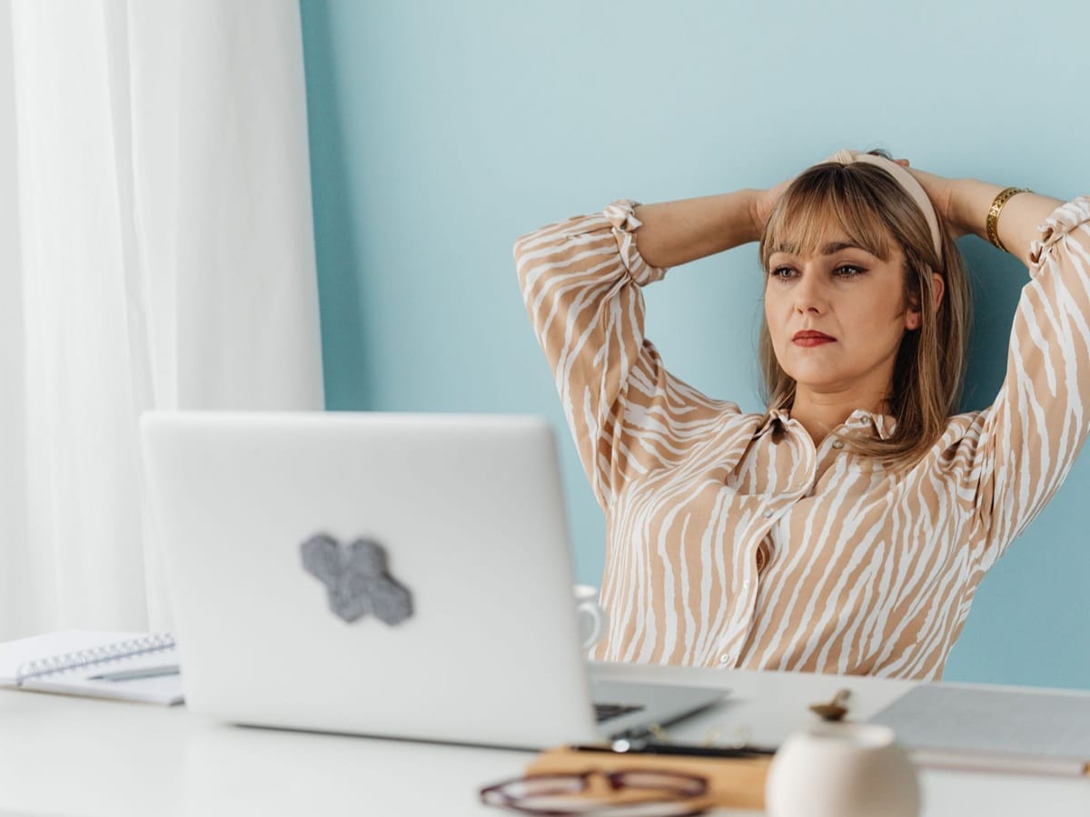 Kobieta siedząca przy laptopie, odchylona do tyłu z rękami za głową, ze skupionym i zamyślonym wyrazem twarzy, zastanawiająca się nad sytuacją w pracy.