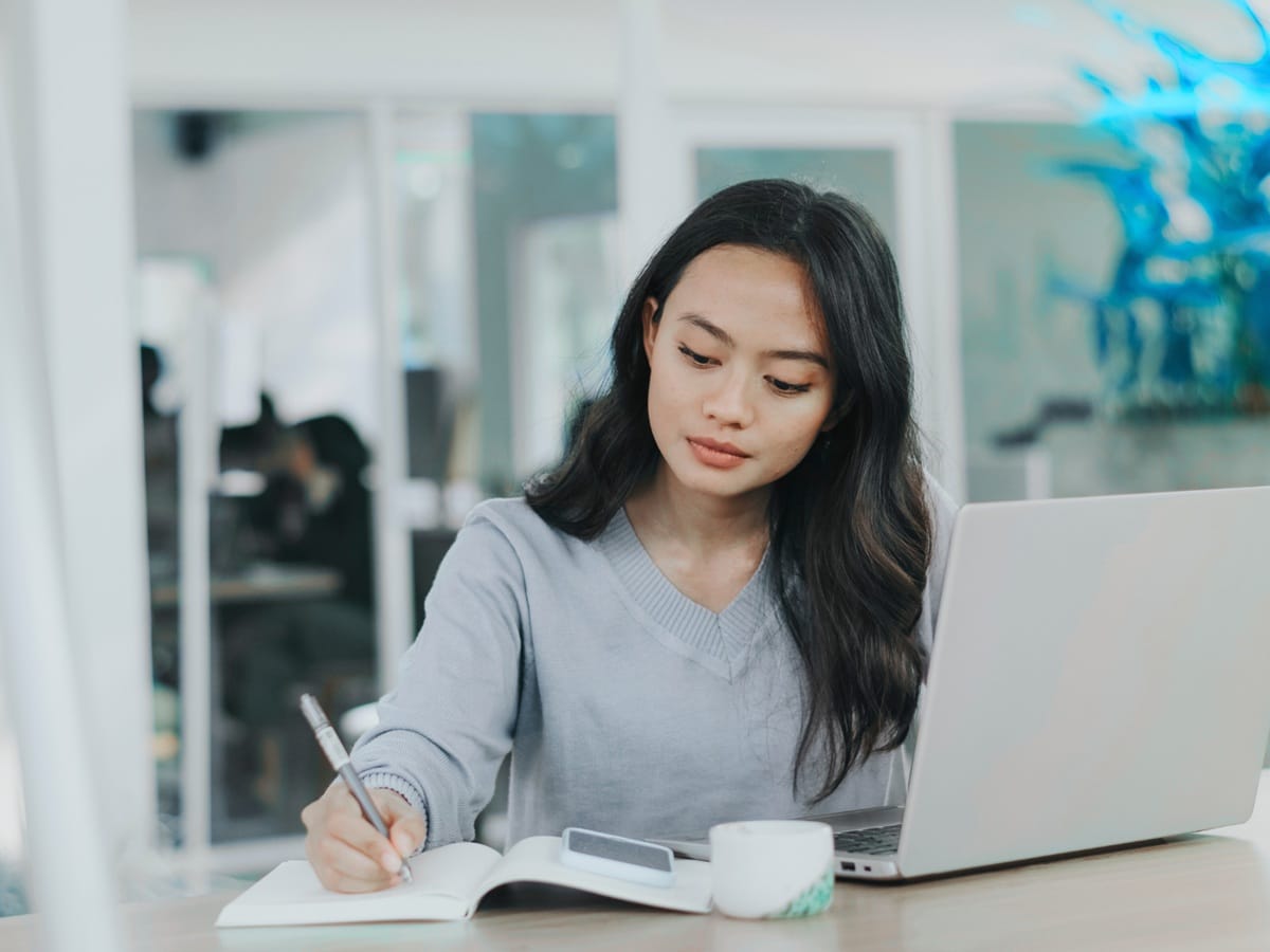 Kobieta pracuje przy laptopie i notuje w skupieniu, symbolizując świadome projektowanie życia i finansów.