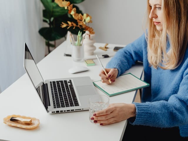 Kobieta planująca finanse w spokojnej przestrzeni, laptop, notatnik, szklanka wody i kadzidełko.