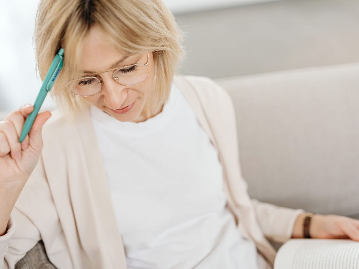 Kobieta siedząca na sofie, zamyślona, z długopisem przy skroni. Przed nią leży otwarta książka, a ona patrzy w bok, jakby przed chwilą przeczytała fragment i zastanawiała się, co zanotować. Naturalne światło, spokojna, minimalistyczna scena, atmosfera refleksji i planowania