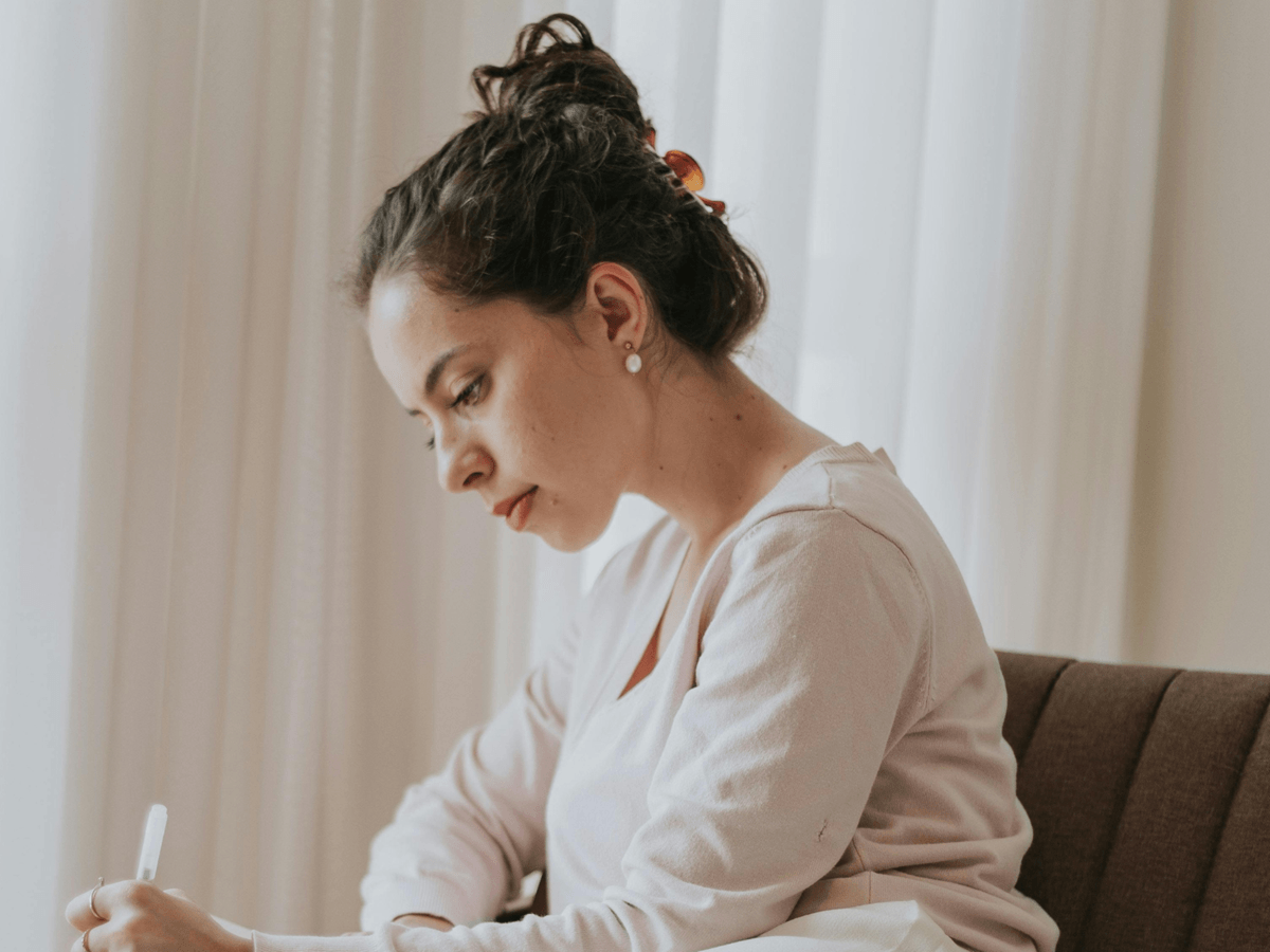 Kobieta siedząca przy oknie, pisząca w skupieniu w notesie. Delikatne światło dzienne, spokojna, refleksyjna atmosfera.
