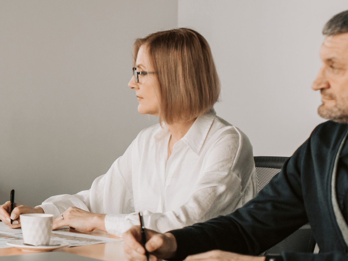 Dwie osoby około pięćdziesiątki siedzą przy stole w skupieniu. Kobieta w białej eleganckiej bluzce słucha w skupieniu, mężczyzna w luźnym garniturze siedzi obok. Oboje patrzą w lewo, słuchając uważnie. Na stole leżą notatki i biała filiżanka, w tle jasna ściana i fragment biznesowego krzesła