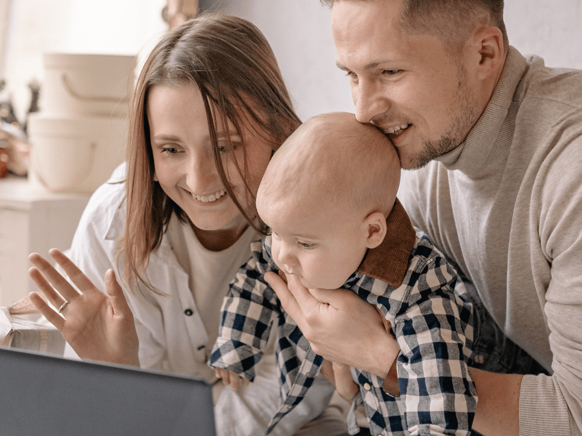 Trzyosobowa rodzina z niemowlęciem siedzi przed laptopem i rozmawia online z bliskimi, pokazując dziecko na ekranie