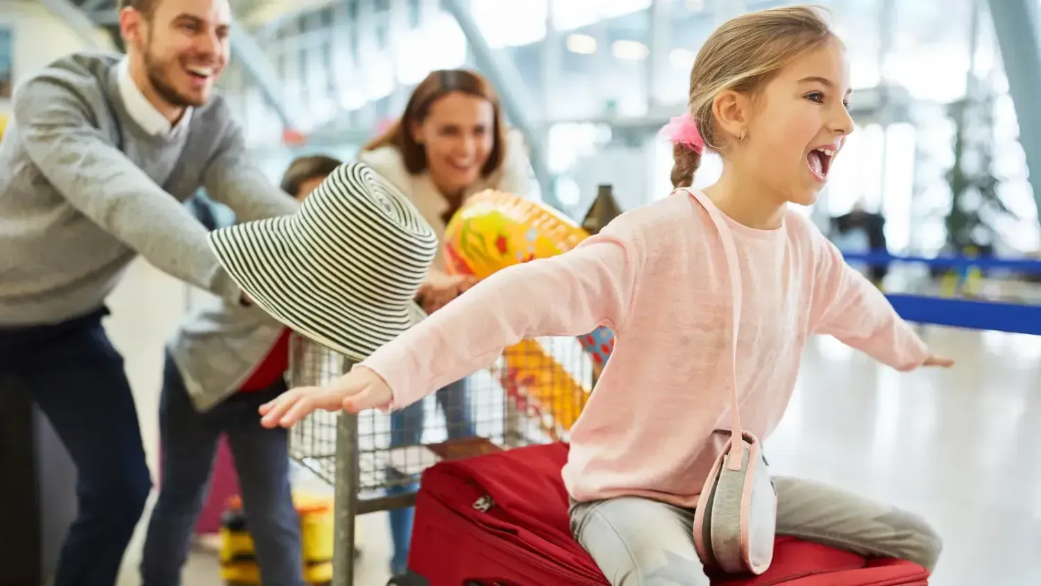 Family at the airport, parents pushing trolley while child rides on luggage with arms outstretched like flying, symbolizing joyful chaos of travel.