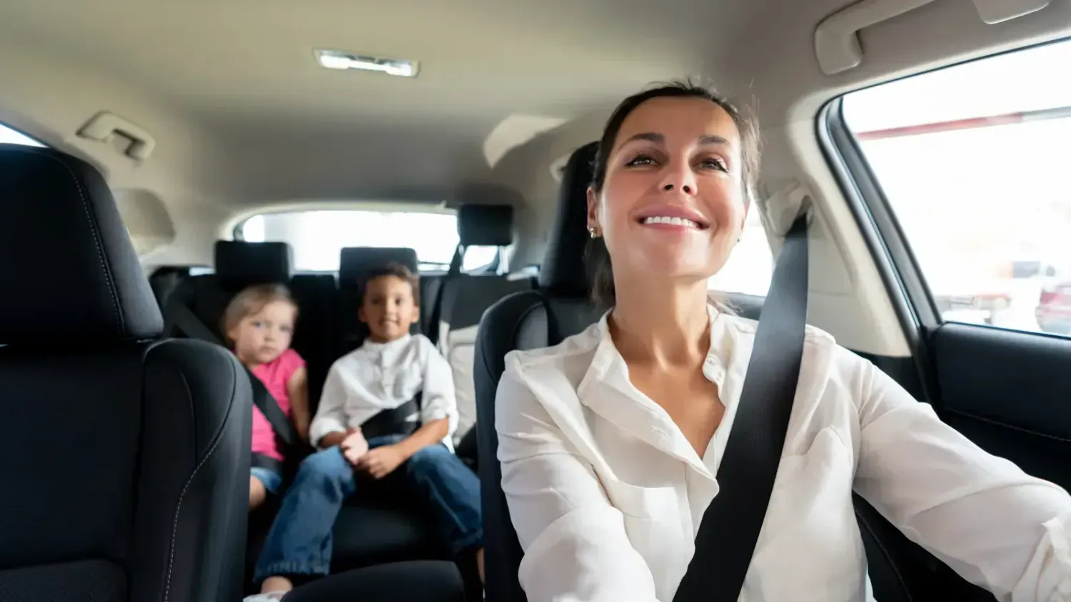 Smiling mom driving with two kids safely buckled in the back seat, representing family car rental and road trip freedom.